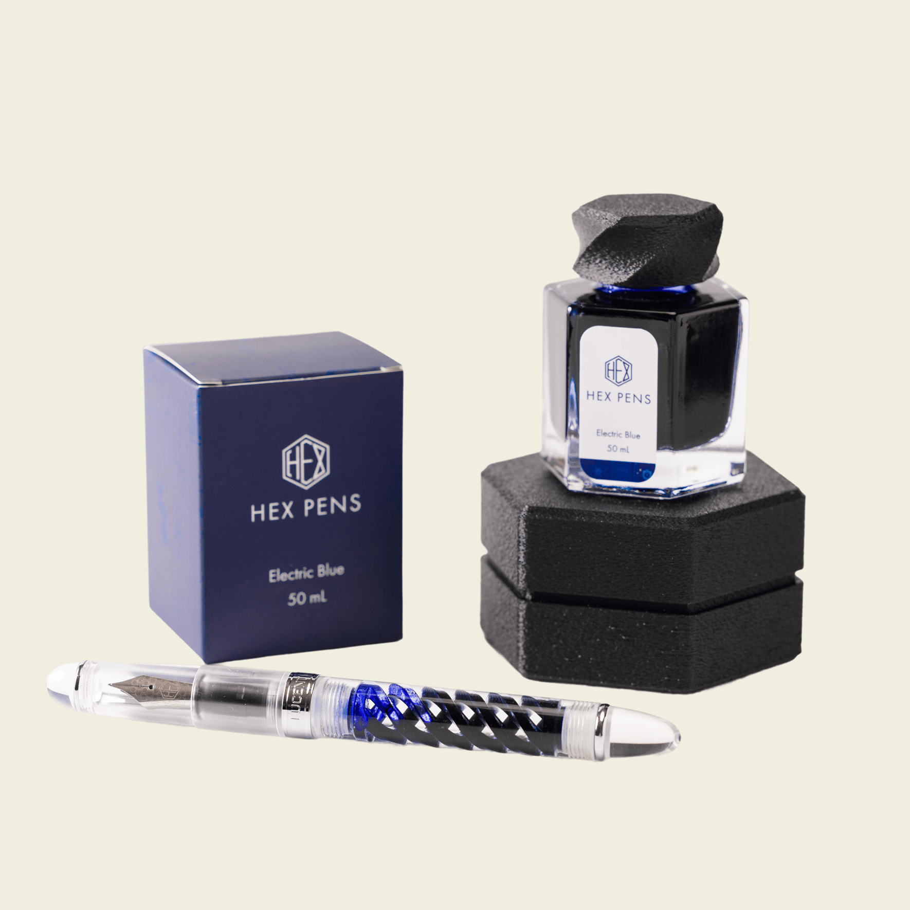 Inked Triple Twist Lucent fountain pen in the front, Electric Blue bottled ink on top of the nib box in the back right, and Electric Blue box in the back left, by Hex Pens.