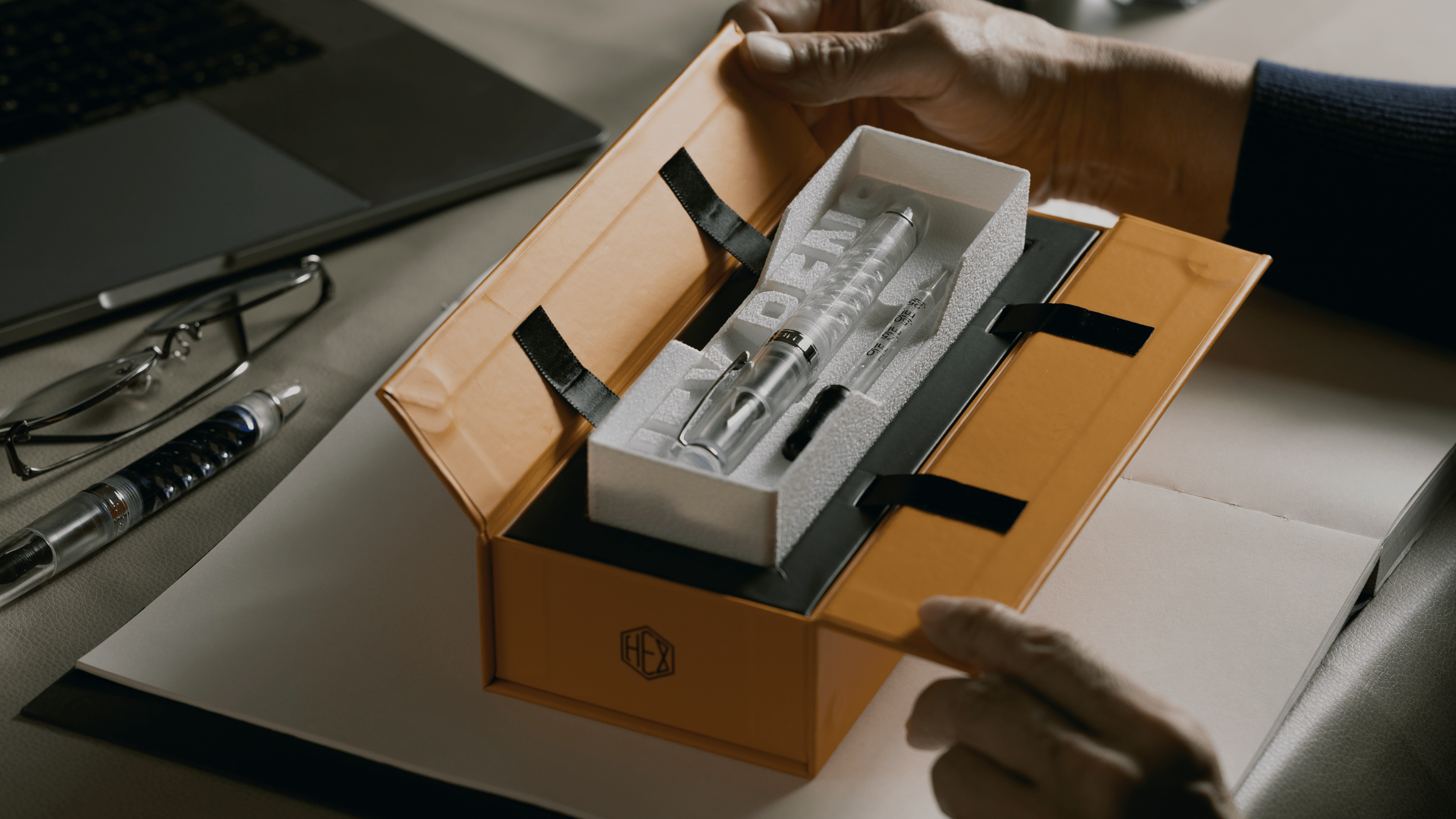Cropped view of uninked Triple Twist Lucent fountain pen with an eyedropper inside the box, both hands in the process of opening it, by Hex Pens.